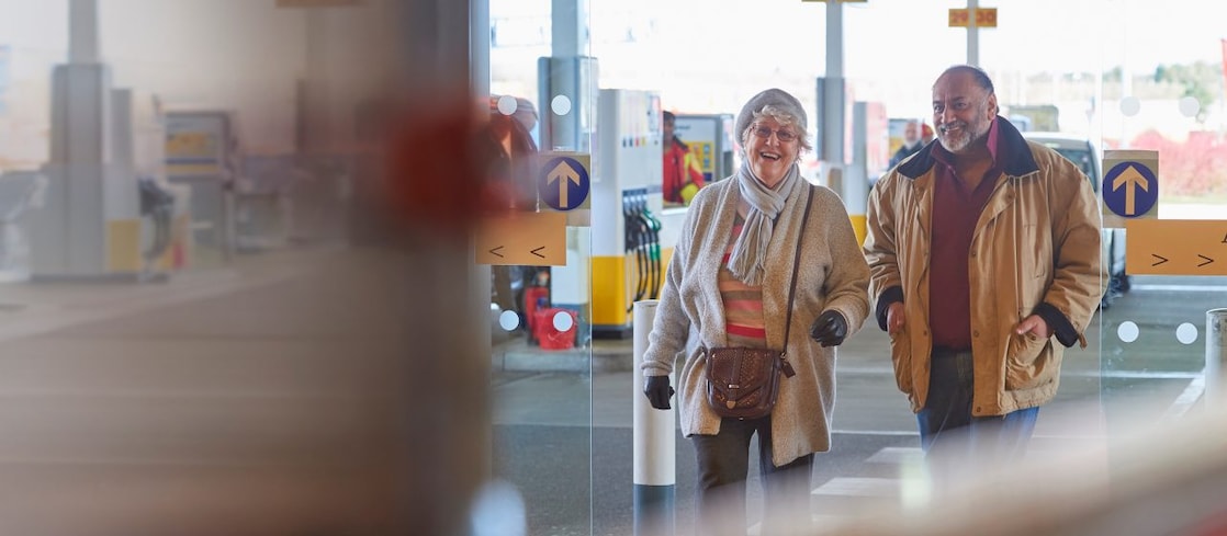 Image shows a lady and man smiling while entering a Shell service station through automatic doors. The image backdrop is a Shell forecourt.