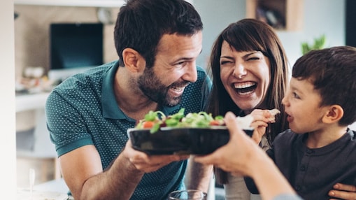 An image of a man, woman and boy hugging and smiling, whilst someone hands them a salad in a black bowl. The blurred background is a kitchen.